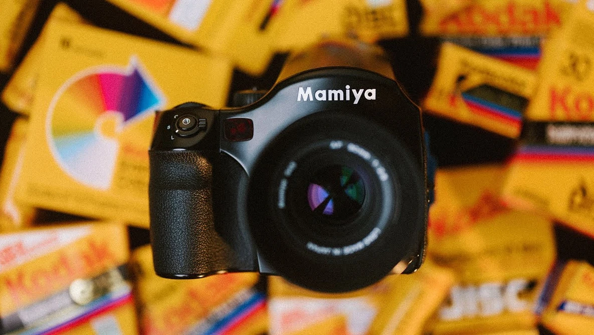 Mamiya medium format camera with colorful film boxes blurred in background.