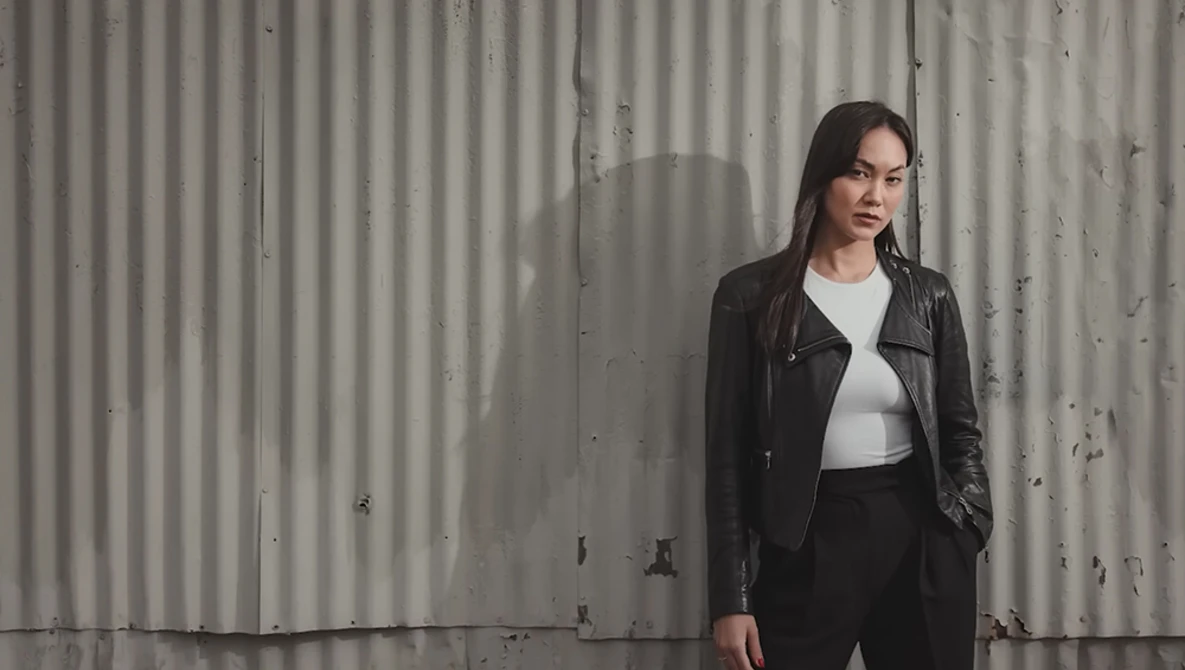 Woman in black leather jacket and white shirt standing against corrugated metal wall.