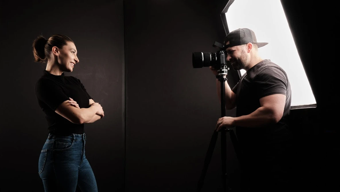 Photographer with camera on tripod shooting portrait of woman in studio with key light.