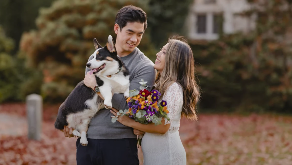Young couple holding a husky dog and fall flowers outdoors in a brick-paved setting.