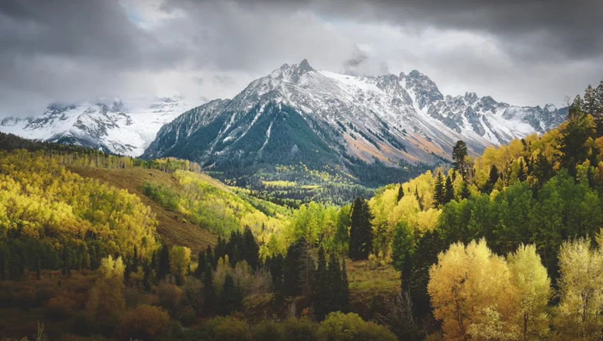 Snow-capped mountain peak rising above a forested valley with golden autumn foliage under dramatic clouds.