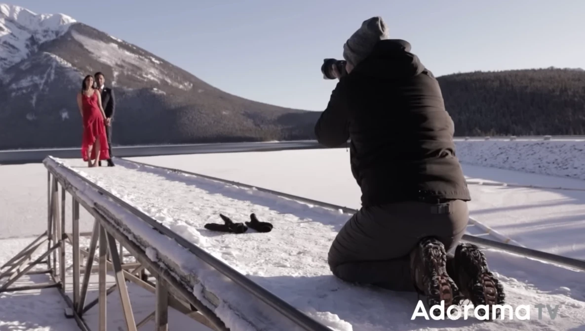 Photographer shooting a portrait of a woman in a red dress standing on a snowy dock, with snow-covered mountains and a frozen lake in the background.