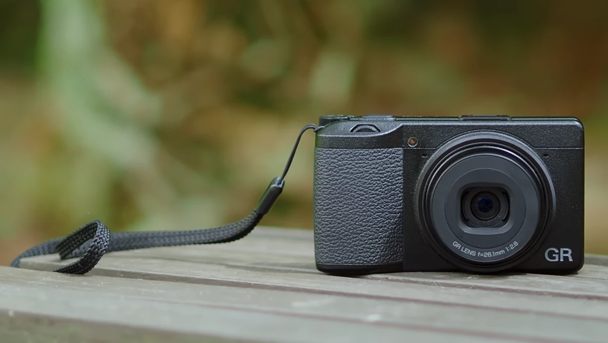 Ricoh GR compact camera with black wrist strap on wooden surface with blurred green foliage background.