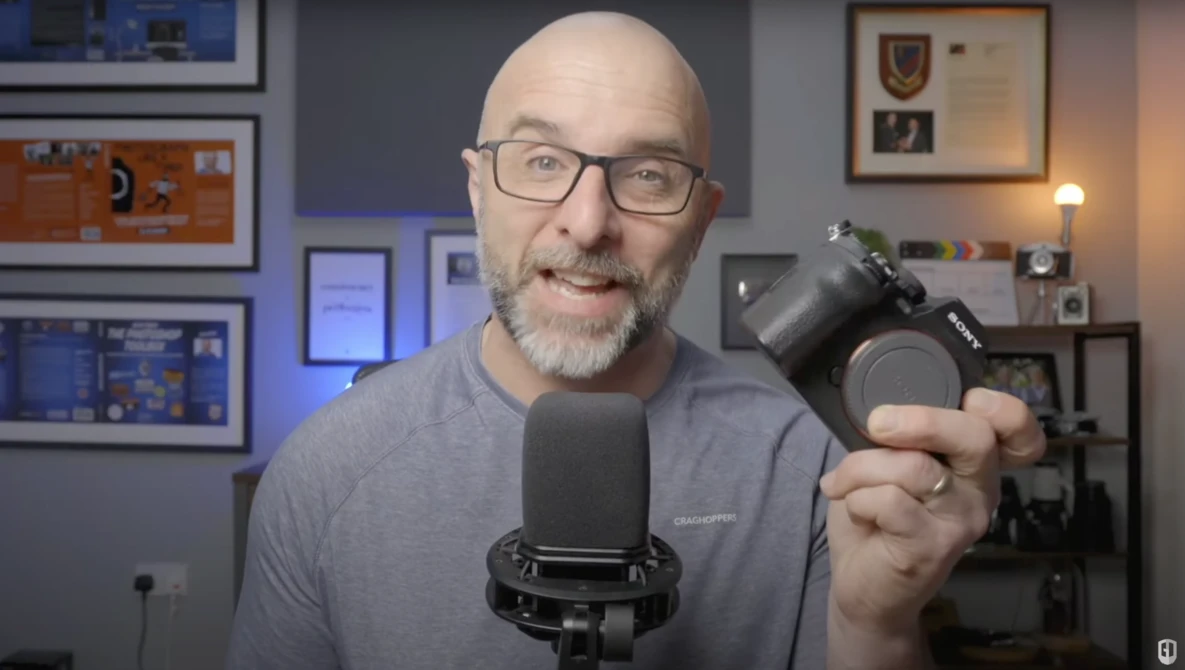 Man holding a camera lens toward the viewer while speaking into a microphone in a studio setting.