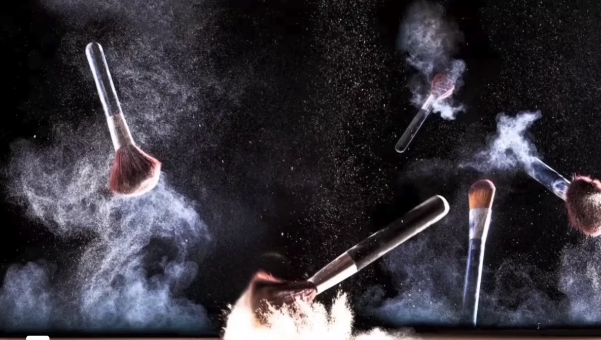 Multiple makeup brushes suspended in mid-air with powder explosions against a dark background.
