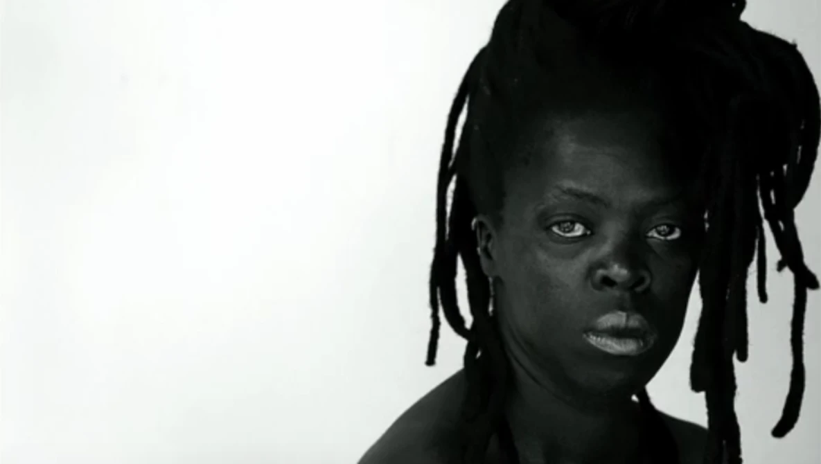Black and white portrait of a person with braided hair looking directly at camera against plain white background.
