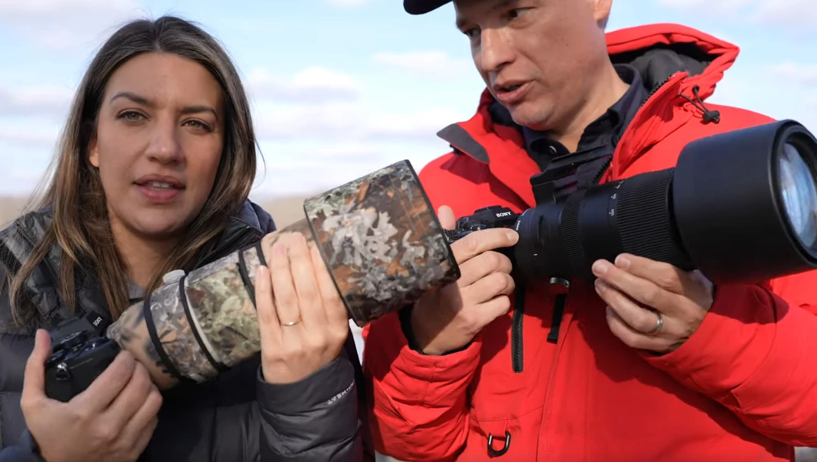 Two photographers holding telephoto lenses with camouflage and red jackets in an open landscape.