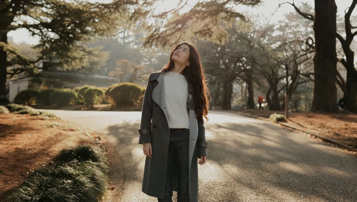 Woman in black coat looking upward while standing on a tree-lined path during golden hour.