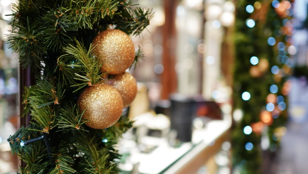 Two gold glitter ornaments hanging from evergreen branch with blurred holiday lights in background.