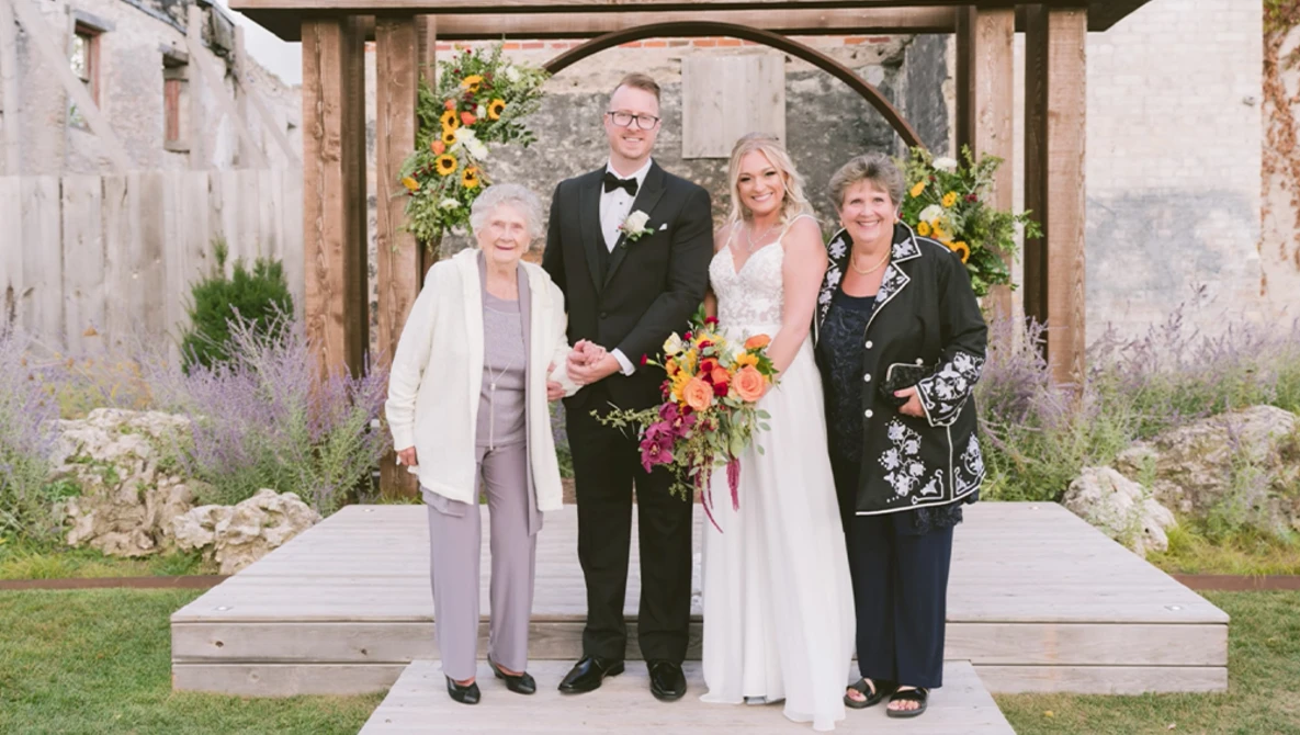Bride and groom pose with two older women under a wooden arch decorated with yellow and pink flowers.