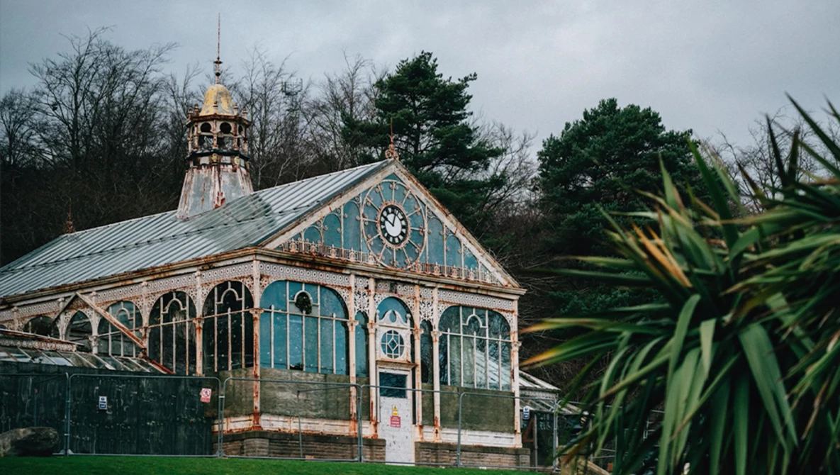 Turquoise Victorian-era glasshouse with ornate cupola and arched windows, flanked by bare trees and evergreens.