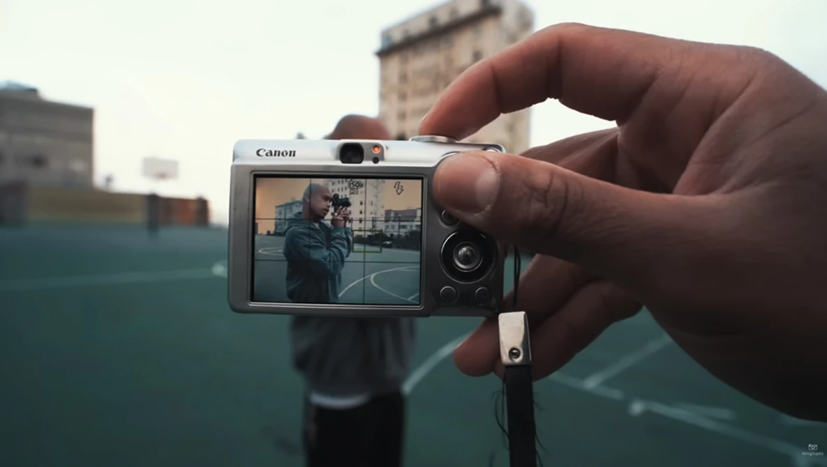 Hand holding a Canon compact camera displaying a street scene on its rear LCD screen.