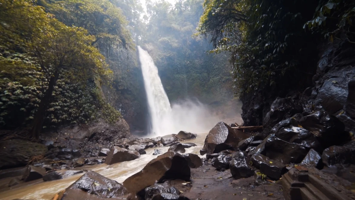 Waterfall cascading down a moss-covered cliff surrounded by dense jungle vegetation and scattered boulders.