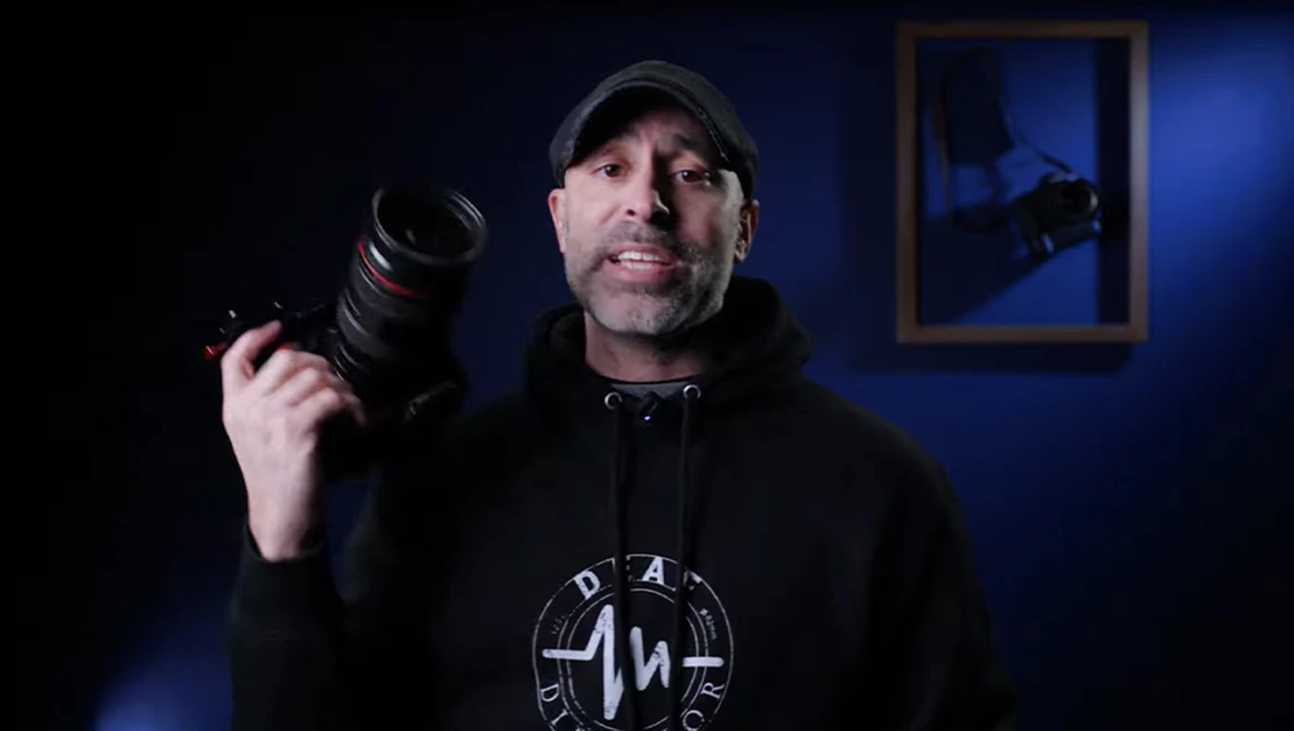 Photographer holding a telephoto lens while wearing a cap and black hoodie against a blue-lit interior backdrop.