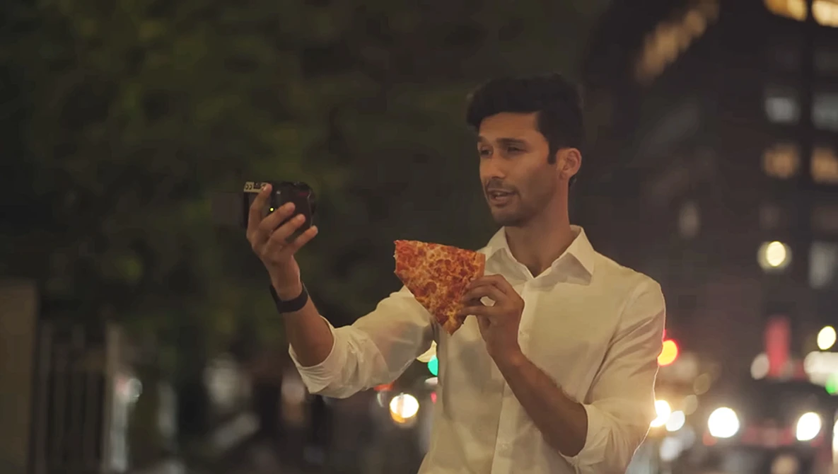 Man in white shirt holding pizza slice while taking selfie on city street at dusk.