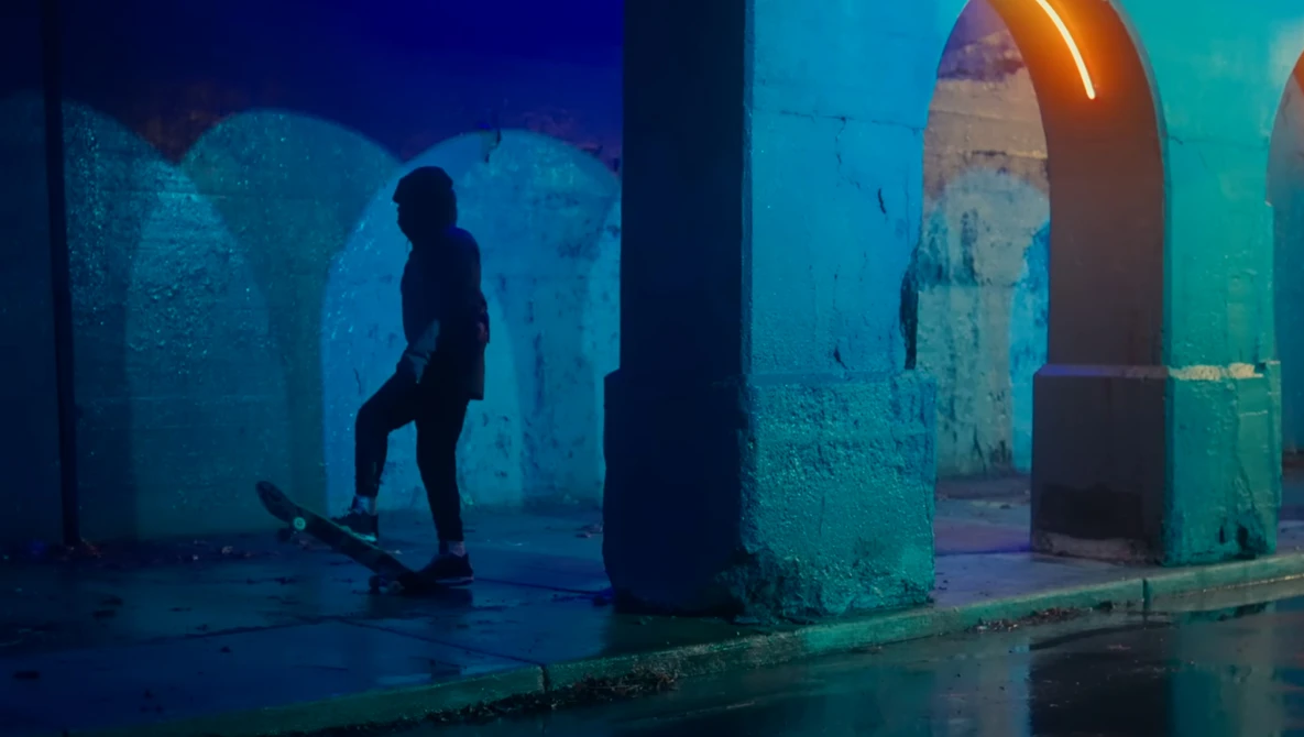 Skateboarder in silhouette under neon blue and teal lighting beneath a concrete overpass.