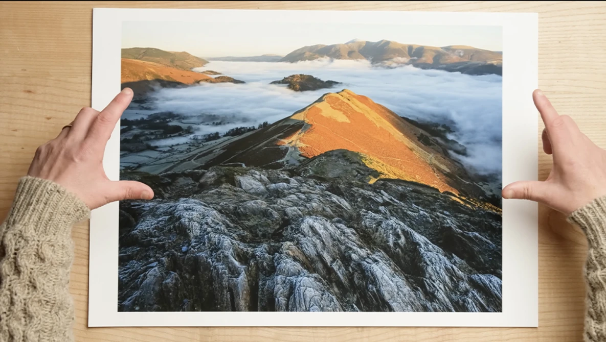 Printed landscape photograph held up against wooden background, showing golden-lit rocky peak above sea-level clouds with islands visible beyond.