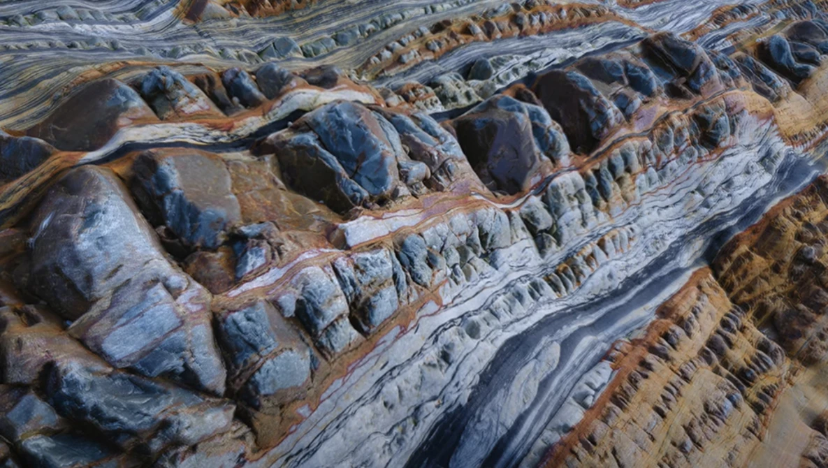 Aerial view of deeply eroded mountain terrain with sharp ridges and layered geological formations in warm earth tones.