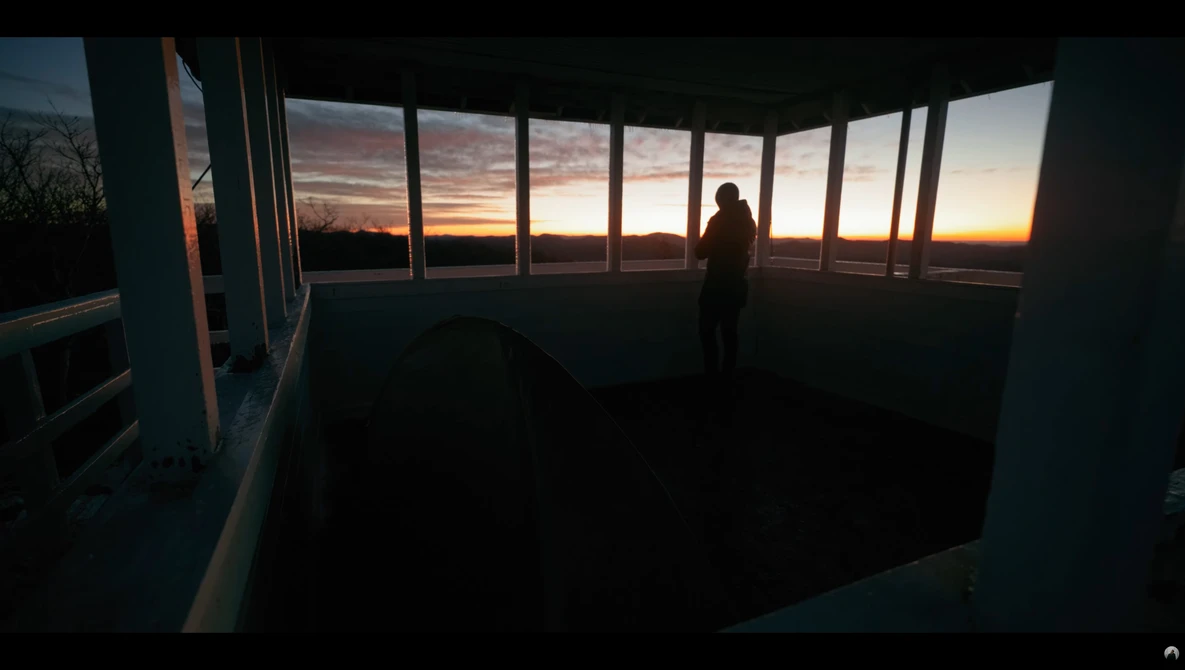 Silhouetted figure standing in a modern glass-walled room overlooking a golden sunset on the horizon.