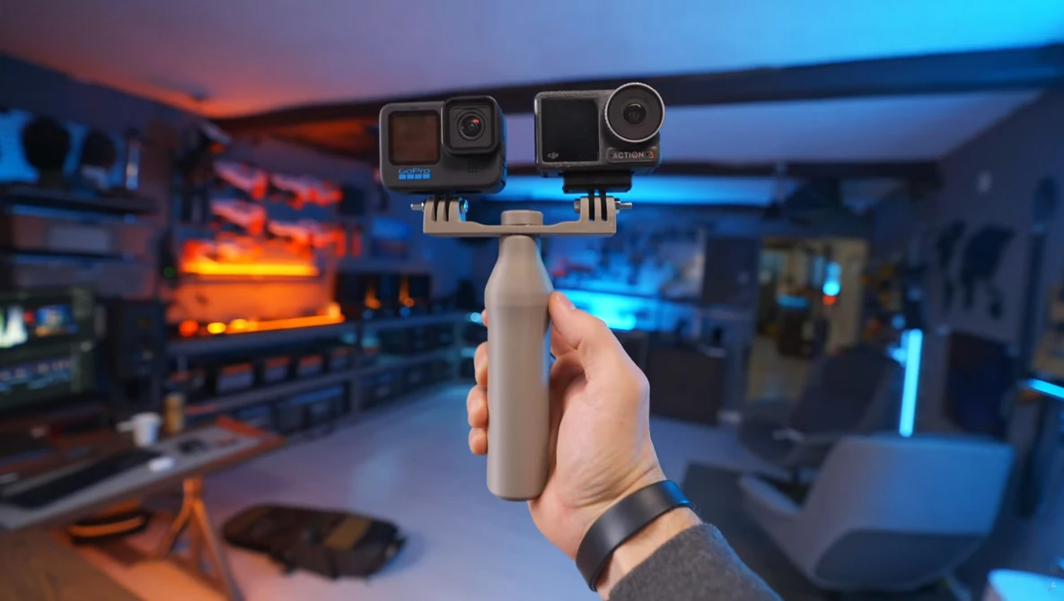 Hand holding a dual camera mounting rig with two action cameras against a blurred tech setup with warm and cool lighting.