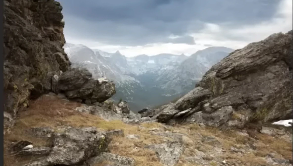 Alpine valley landscape framed by rocky peaks with dramatic cloudy sky and distant mountains.