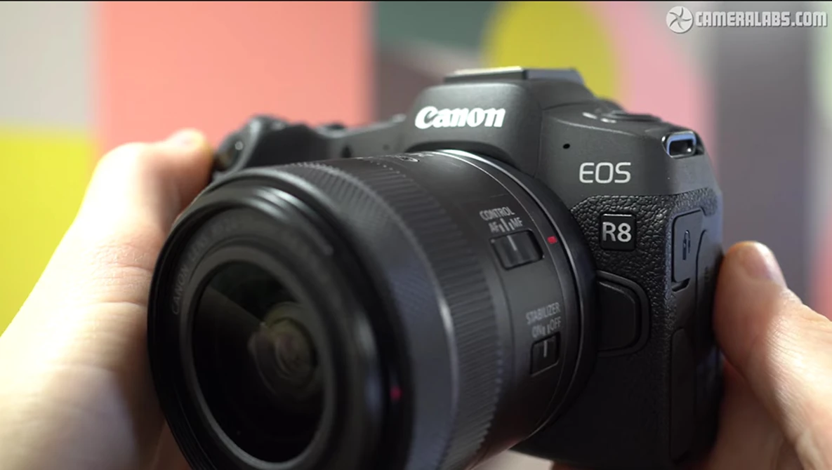 Canon EOS R8 mirrorless camera held in hands with attached lens against colorful background.