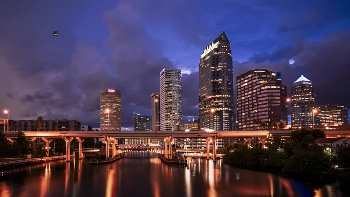 Illuminated downtown skyline reflected in a river at dusk, with a bridge spanning the water.
