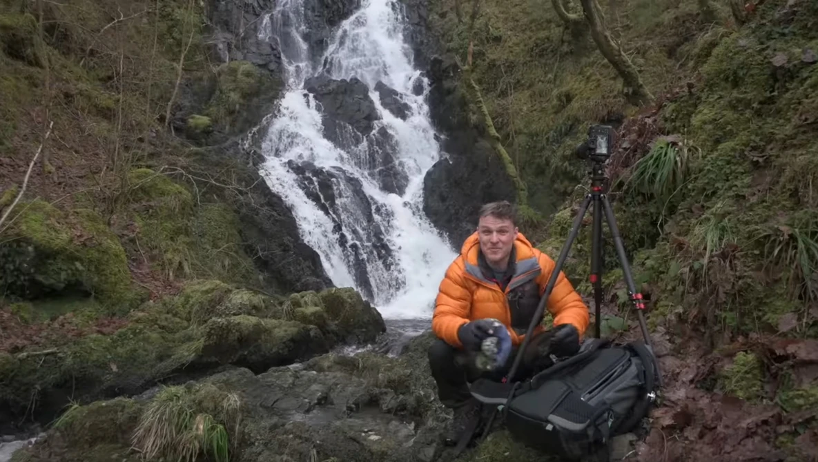 Photographer in orange jacket crouching by waterfall with camera gear in forested landscape.