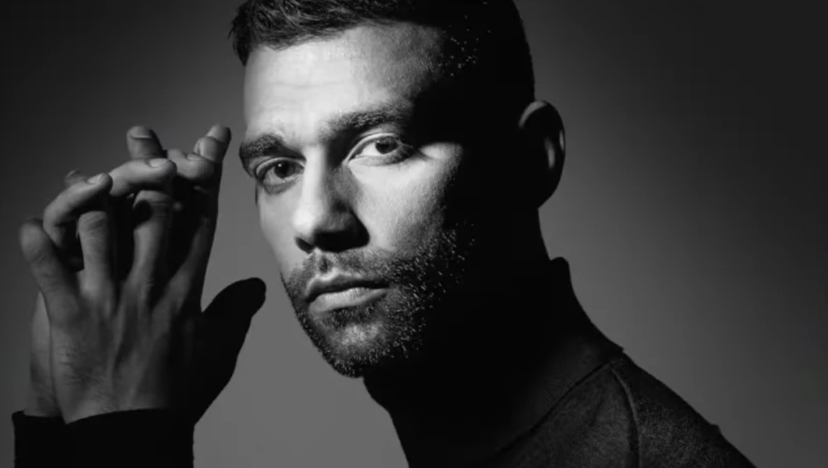 Black and white portrait of a bearded man with hand raised to his face, studio lighting against neutral background.