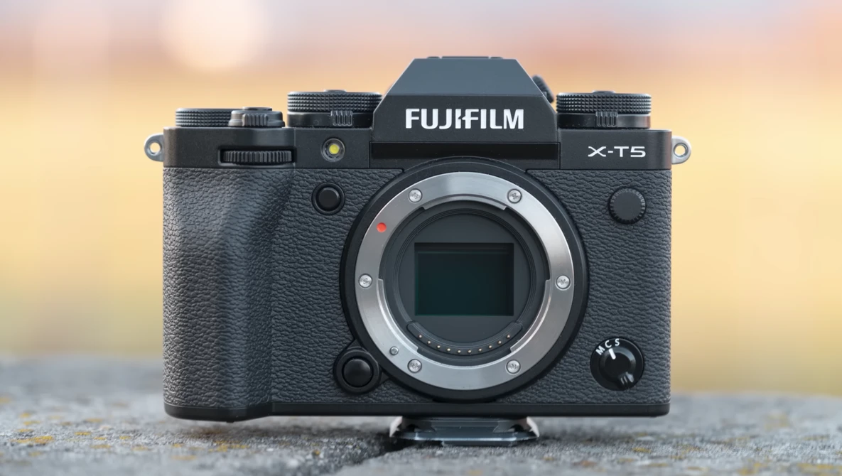 Fujifilm X-T5 mirrorless camera body with lens mounted, displayed front-facing against a blurred warm-toned background.