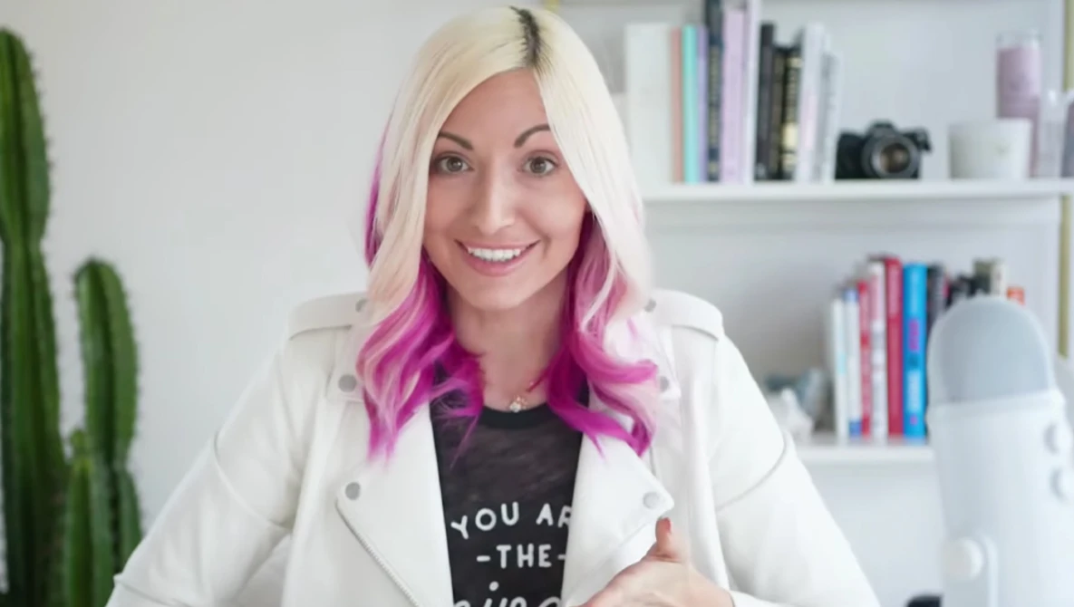 Woman with blonde and magenta hair wearing white blazer and black graphic tee smiles at camera in home office setting.