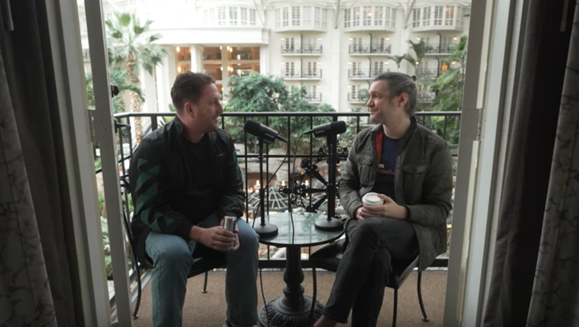 Two men having a conversation on a balcony overlooking a European-style urban streetscape.