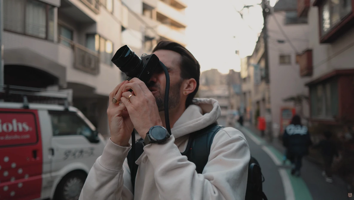 Male photographer holding camera up to his face on an urban street lined with buildings.