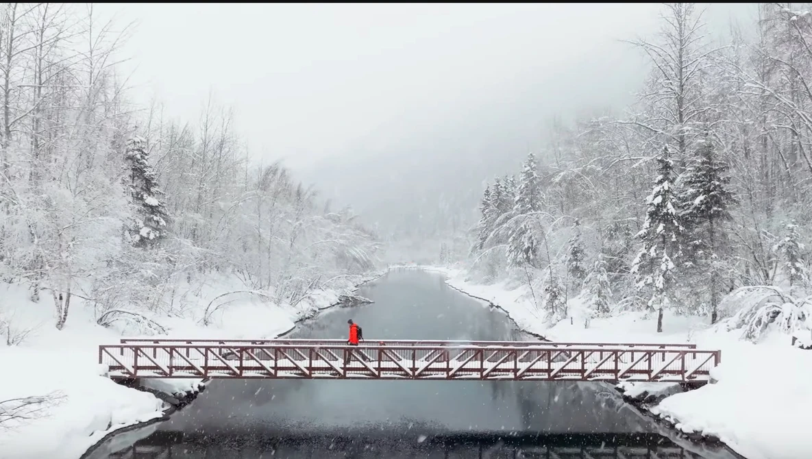 Person in red jacket standing on wooden bridge over partially frozen river surrounded by snow-covered forest.