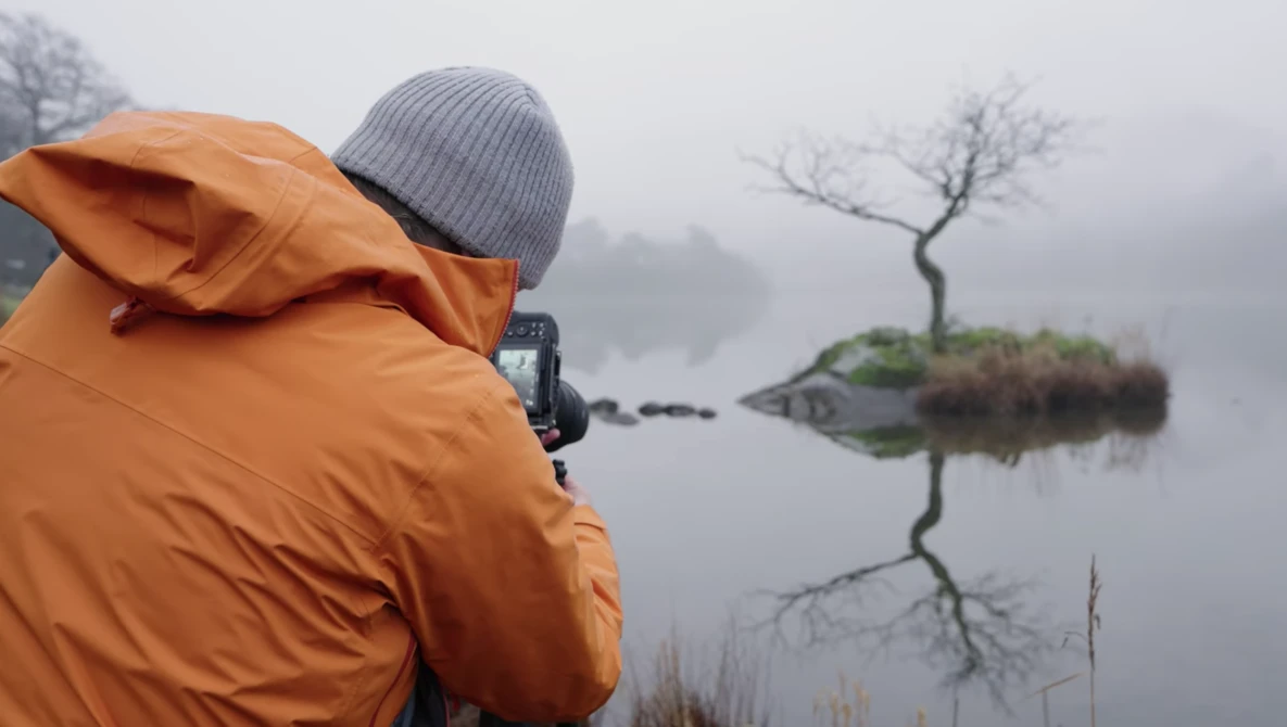 Photographer in orange jacket shooting a minimalist landscape with a solitary tree reflected in still water on a foggy morning.