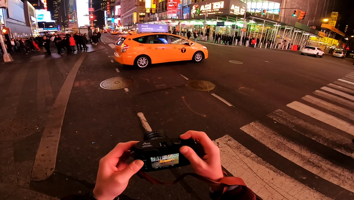 Hands holding a smartphone capturing an orange taxi cab at a busy city intersection at night.
