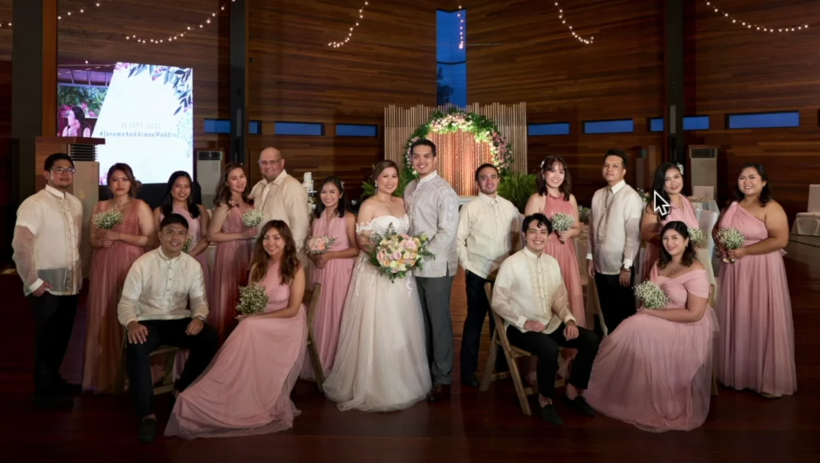 Wedding party of 18 people in coordinated pink and cream attire posed together in a wooden chapel.