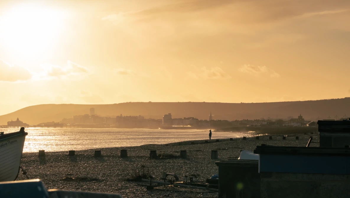 Golden hour light illuminates a coastal town across the water from a rocky beach with moored boats in the foreground.