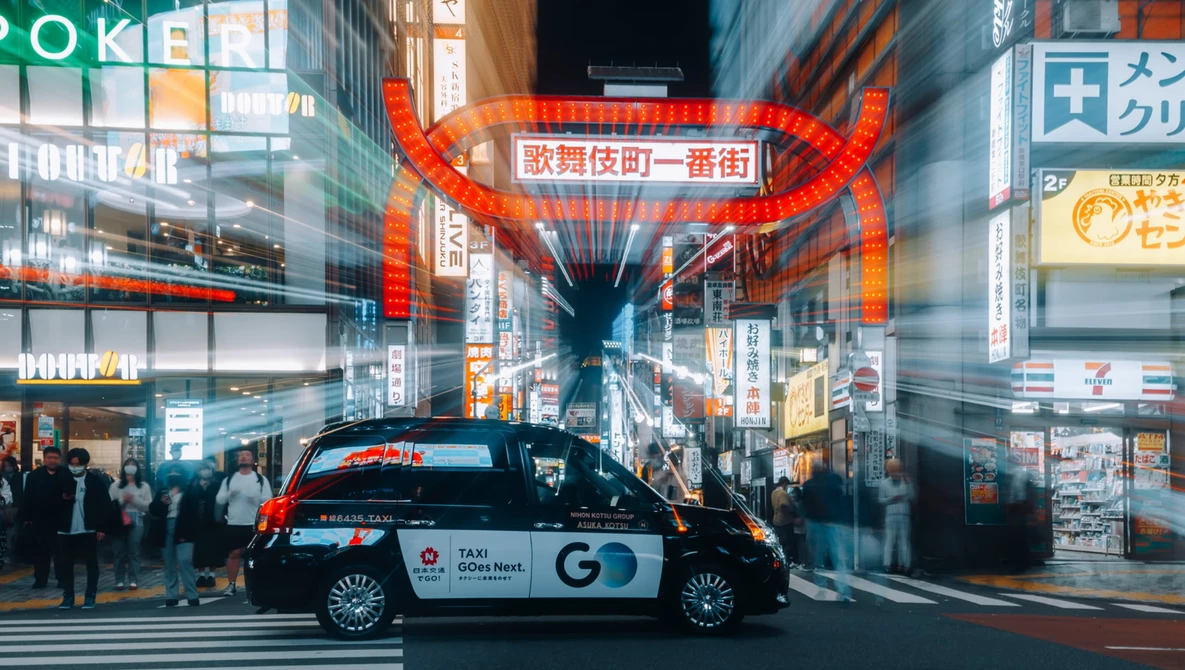 Taxi parked in busy Japanese street intersection at night with large red neon signage overhead.
