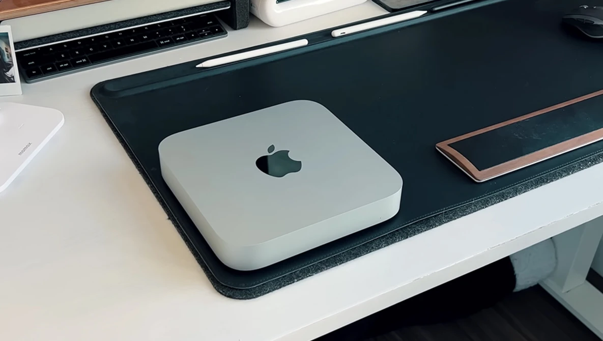 Mac mini computer on a black desk pad next to a keyboard and other workspace items.