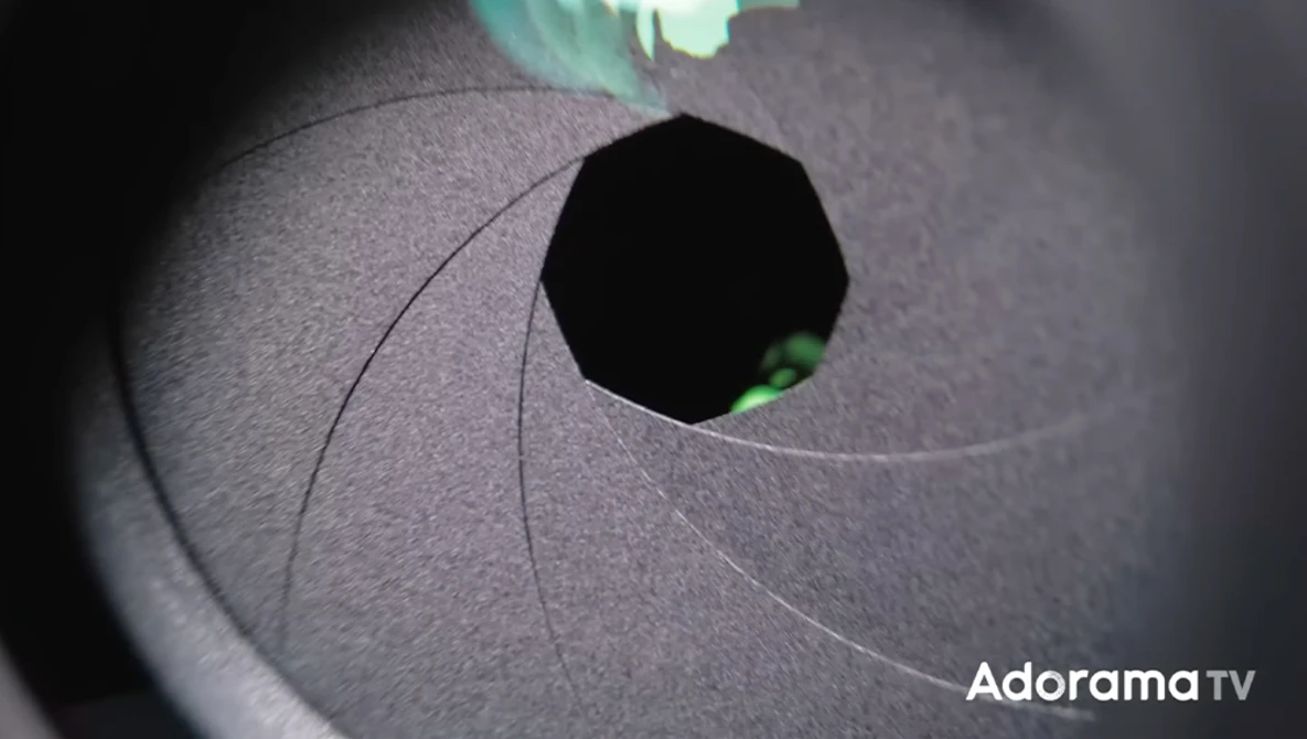 Close-up of a camera aperture blade assembly with a bright green eye visible through the opening.