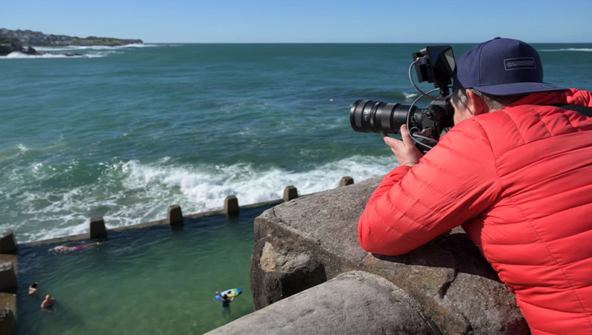 Photographer in red jacket shooting ocean waves from rocky outcrop with telephoto lens.