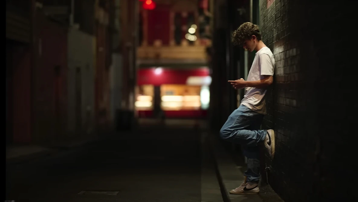 Man leaning against brick wall at night checking mobile phone on dimly lit urban street.
