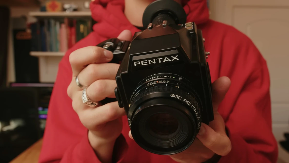 Person in red shirt holding a black Pentax film camera with SMC Pentax lens toward the camera.