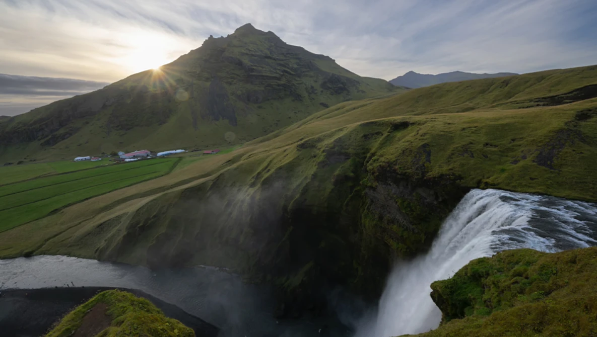 Powerful waterfall cascades down moss-covered cliffs in a verdant valley beneath a distinctive peaked mountain.