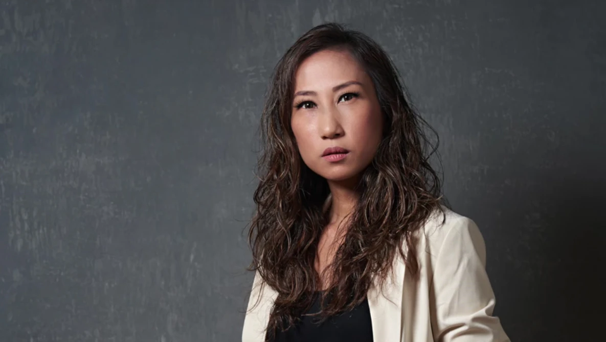 Woman with long dark wavy hair wearing a light beige cardigan against a textured gray backdrop.