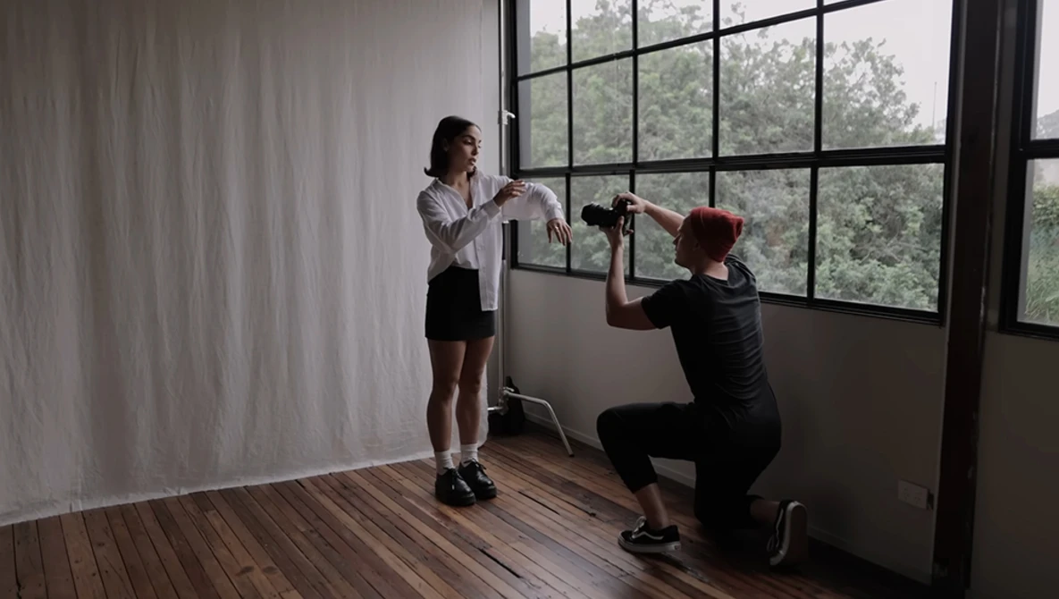 Photographer crouching to shoot while model poses by large windows overlooking trees.