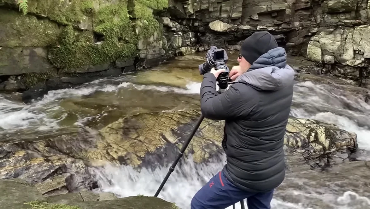 Photographer in black jacket holding camera on selfie stick while shooting a flowing stream surrounded by moss-covered rocks.