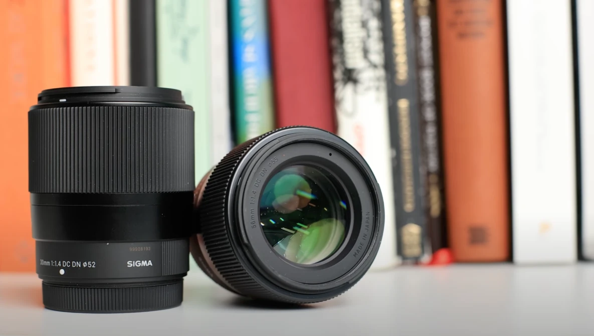 Two Sigma camera lenses displayed on a surface with colorful books in soft focus behind.