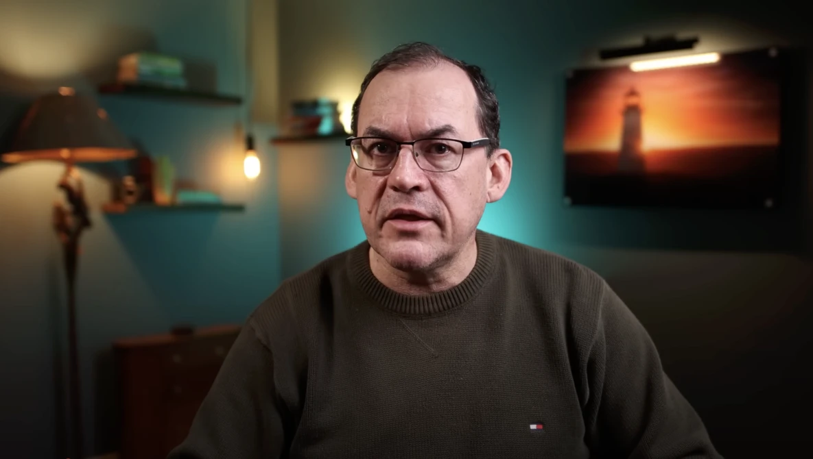 Man wearing glasses and dark sweater speaking in a studio setting with teal walls and ambient lighting.